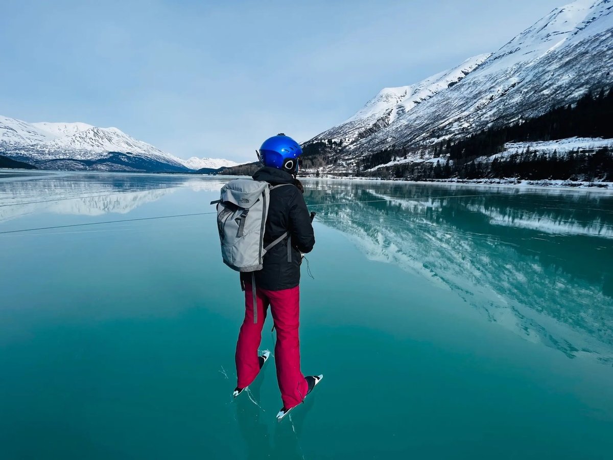 an ice skater stands on very clear blue lake ice with mountains in the background