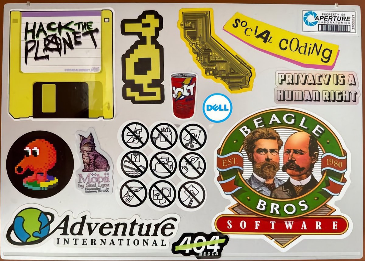 Laptop sticker collection featuring gaming and tech references including Pac-Man, pixel art, Aperture Science, Dell, and various programming-related stickers.