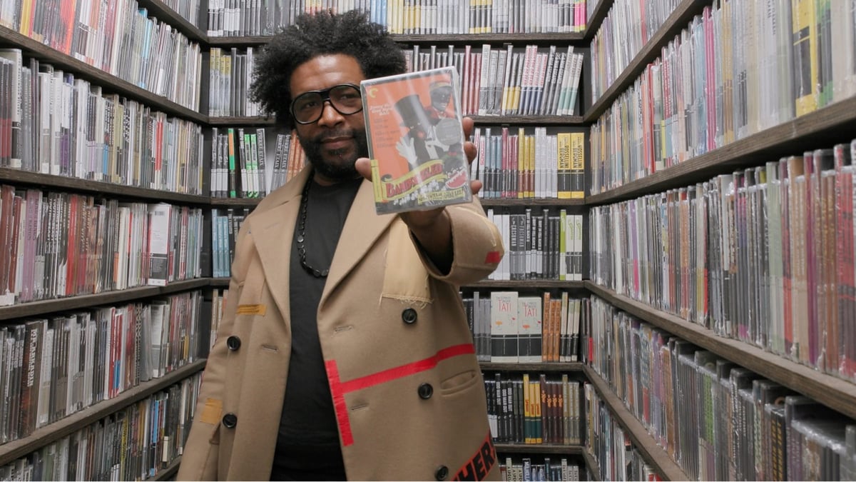 Questlove in the Criterion closet holding up a DVD surrounded by shelves of hundreds of movie DVDs