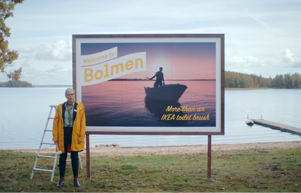 a woman standing in front of a billboard that reads 'Welcome to Bolman, more than an IKEA toilet brush