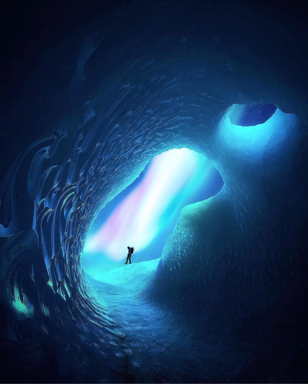 a lone figure stands silhouetted in the entrance of a blue ice cave with the northern lights behind them